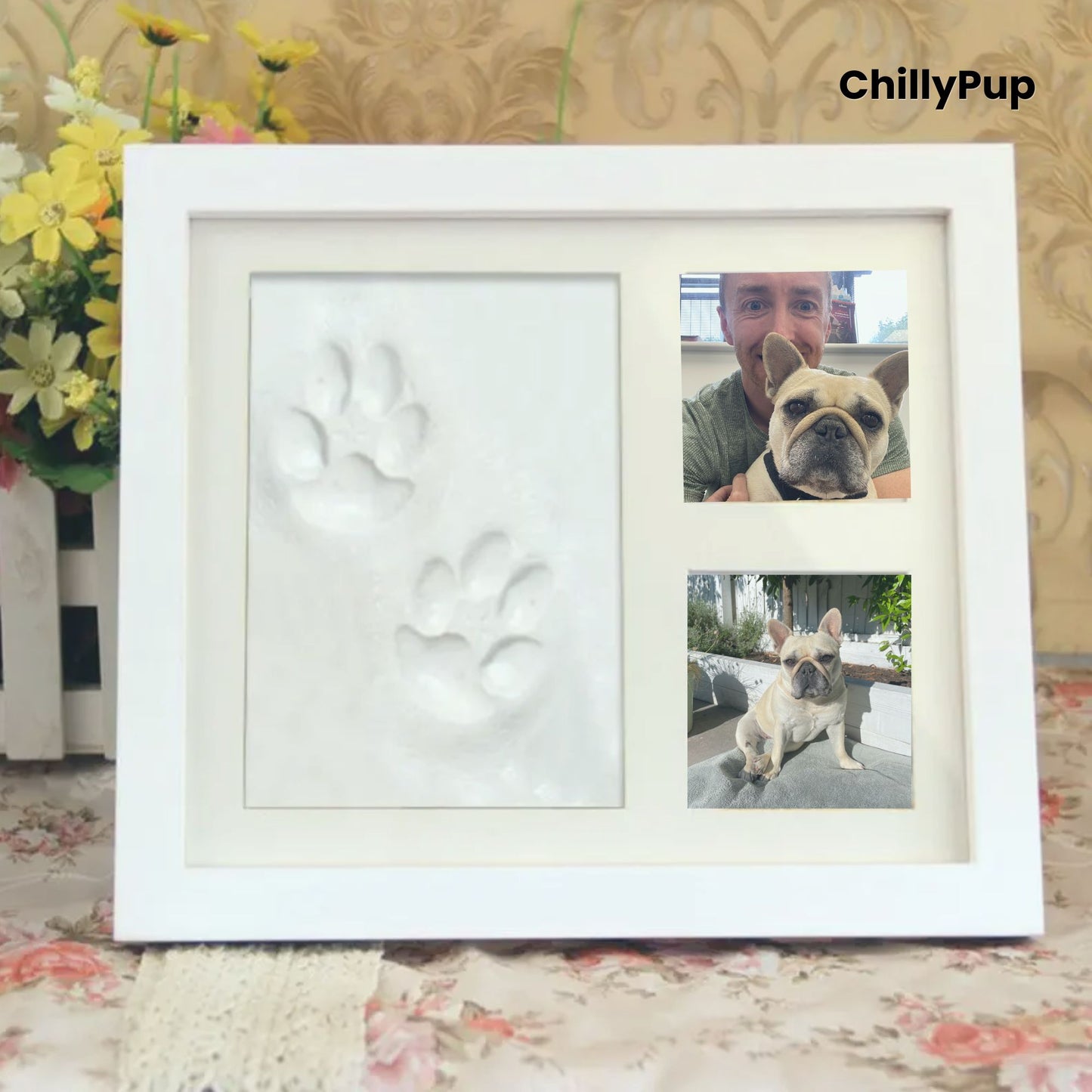 White photo frame with paw print and two pictures of a dog, on a floral surface with flowers in the background.