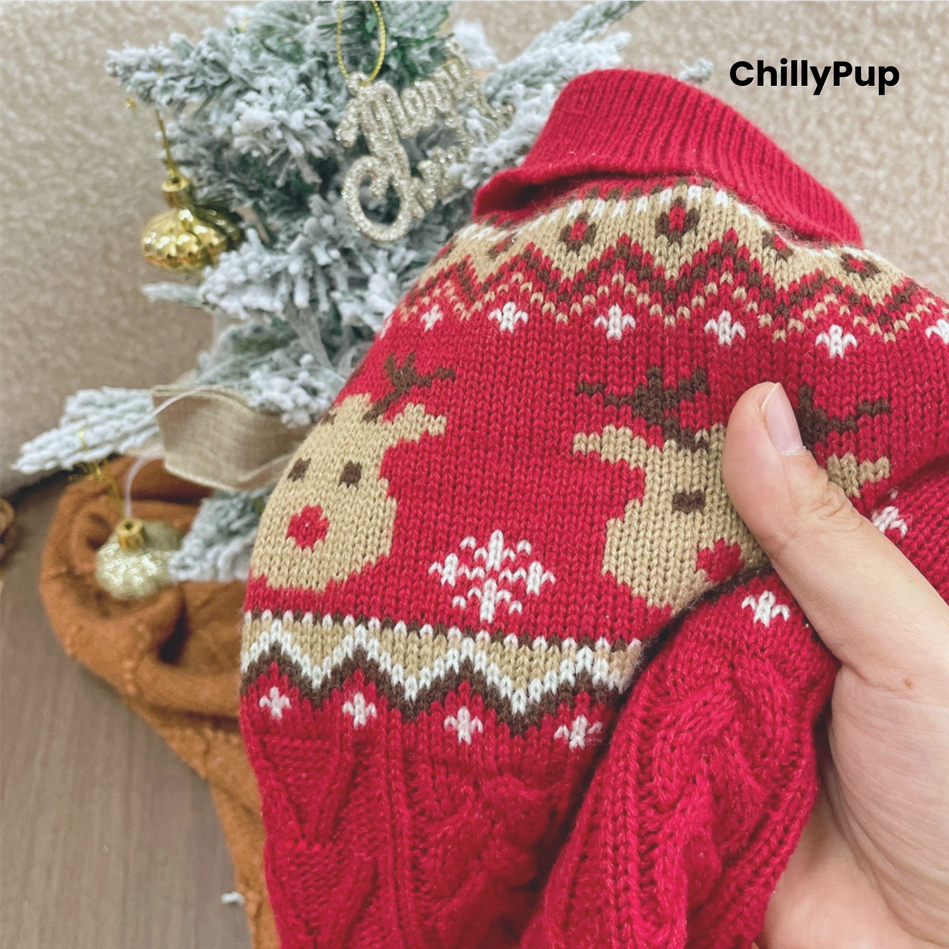 Red knitted sweater with reindeer pattern held by a hand, Christmas tree in the background